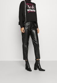 Pull noir court avec le texte « BRAVE LJ WOMAN », associé à un pantalon en cuir noir taille haute et des bottines, avec un design à bout carré.