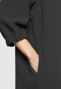 Prenda negra de corte holgado, con mangas abullonadas y bolsillos laterales. La tela es suave al tacto, con una textura delicada y un brillo sutil.