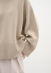 Beige pletený svetr s žebrovaným lemem a zápěstími; má volný střih a měkkou texturu, v kombinaci s světlými, volnými kalhotami.