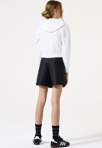Weißer, flauschiger Crop-Hoodie, schwarze Shorts aus Kunstleder, schwarze Socken mit Text und schwarze Sneaker mit weißen Streifen. Neutraler Hintergrund.