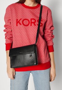 Felpa rossa con motivo del logo ripetuto e polsini a costine, abbinata a una borsa a tracolla in pelle nera con pattina e chiusura in argento.
