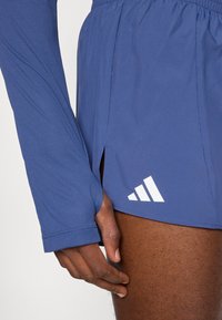 Μπλε αθλητικό σορτς με πλαϊνή σχισμή και λευκό λογότυπο Adidas στο τελείωμα. Το ύφασμα φαίνεται ελαφρύ και λείο, κοντά στο δέρμα.