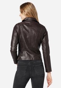 Bruine leren jas met een aansluitend ontwerp, geknoopte kraag en naaddetails, gecombineerd met zwarte jeans, in de achteraanzicht.