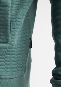 Sudadera con capucha verde texturizada con un tejido estampado, mangas lisas y una pequeña etiqueta negra con "PETROL IND" en el lateral.