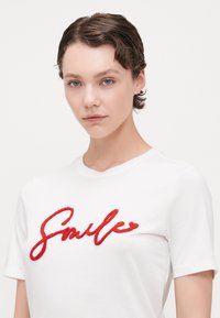 Hvit bomull t-skjorte med rød, teksturert "Smile" skrift over brystet. Korte ermer og klassisk rund halsdesign.