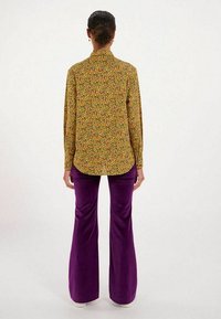 Blouse à manches longues, col, de couleur jaune avec un motif floral multicolore, associée à un pantalon en velours violet, taille haute et évasé.
