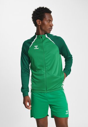 Mann in grüner Sportjacke und Shorts steht mit einer Hand in der Tasche und sieht zur Seite vor einem schlichten weißen Hintergrund.