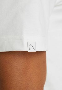 Weißes Baumwoll-T-Shirt mit sichtbarer Textur im Stoff. Es verfügt über ein kleines Label, das am Ärmel angenäht ist und ein einfaches schwarzes Grafikdesign zeigt.