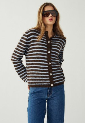 Frau trägt übergroße schwarze Sonnenbrille, eine braun-hellblau gestreifte, geknöpfte Strickjacke und blaue Jeans, posiert vor einem einfarbigen Hintergrund.