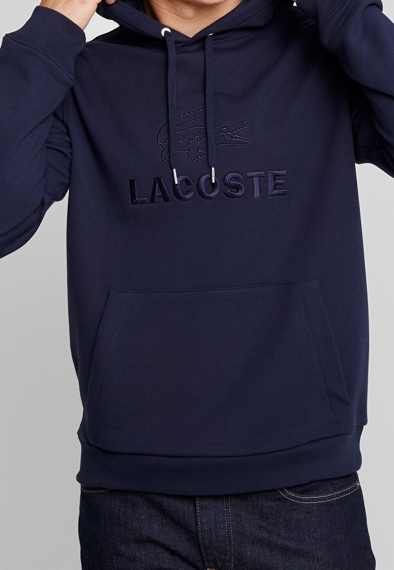 Námořnický modrý mikina s klokaní kapsou, šňůrkami a výšivkou loga Lacoste. Měkký materiál, klasický design a žebrované manžety.