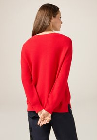 Czerwona sweter z miękką teksturą, długimi rękawami i szerokim dekoltem. Ma prosty krój i boczne rozcięcia, noszony z ciemnoniebieskimi spodniami dżinsowymi.