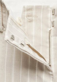 Gros plan sur un pantalon beige à rayures blanches verticales, montrant une fermeture éclair, un bouton et des agrafes partiellement ouvertes.