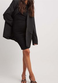 Schwarzes Bodycon-Kleid mit Raffung, kombiniert mit einem übergroßen schwarzen Blazer. Das Model trägt schwarze Riemchenheels und zeigt eine glatte Beintextur.