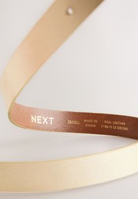 Cintura in vera pelle color oro con finitura liscia. L'interno presenta un'etichetta marrone scuro con la scritta "NEXT SMALL MADE IN CHINA REAL LEATHER".