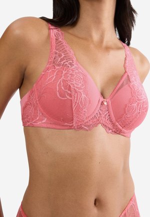 Kvinde iført pink blonder-bh med blomsterbroderi og brede stropper, vist fra skuldre til talje mod hvid baggrund.