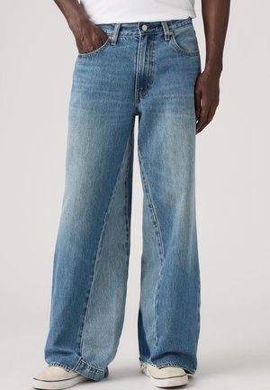 Personne portant un jean bleu large, clair et moyen, les mains dans les poches et des baskets blanches sur fond uni.