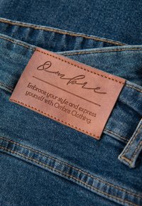 Dunkelblauer Denim mit einem aufgenähten braunen Lederetikett. Das Etikett zeigt geprägten Text, einschließlich einer Unterschrift und einer Markenbotschaft.