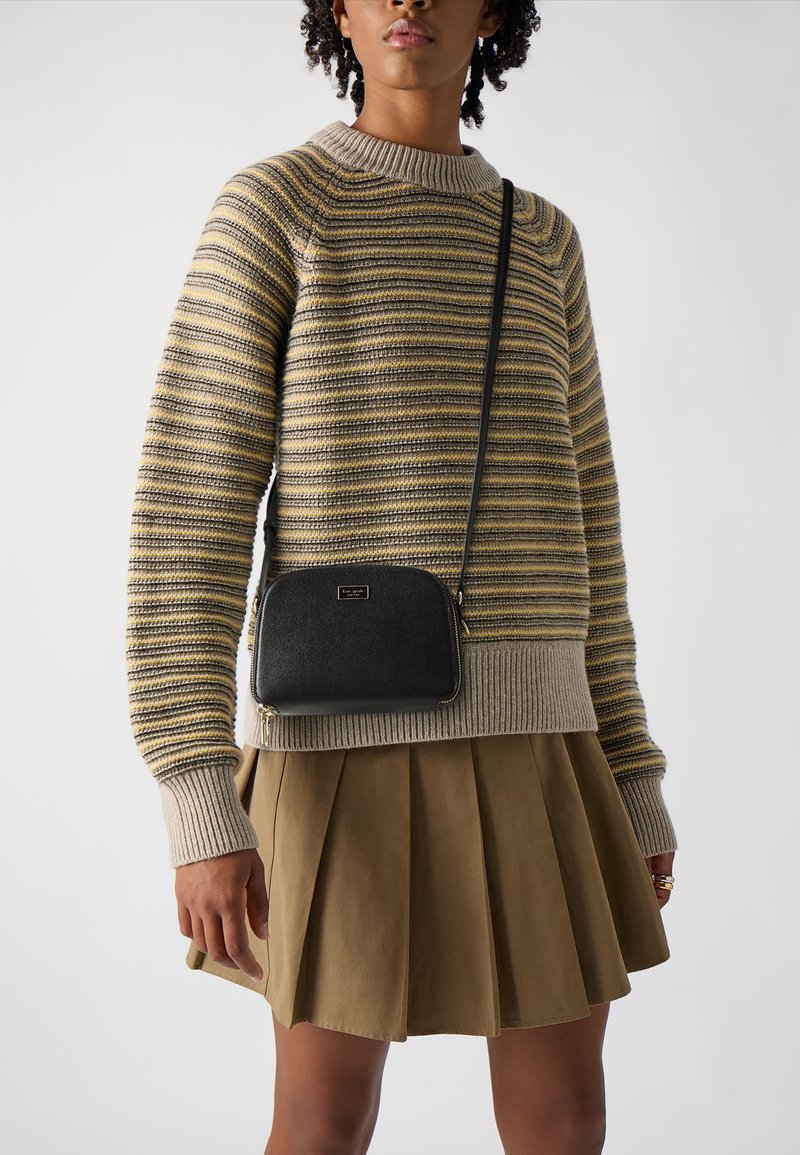 Una borsa a tracolla nera con forma arrotondata, texture liscia, zipper dorato e logo del brand, abbinata a un maglione a righe in maglia e a una gonna plissè.