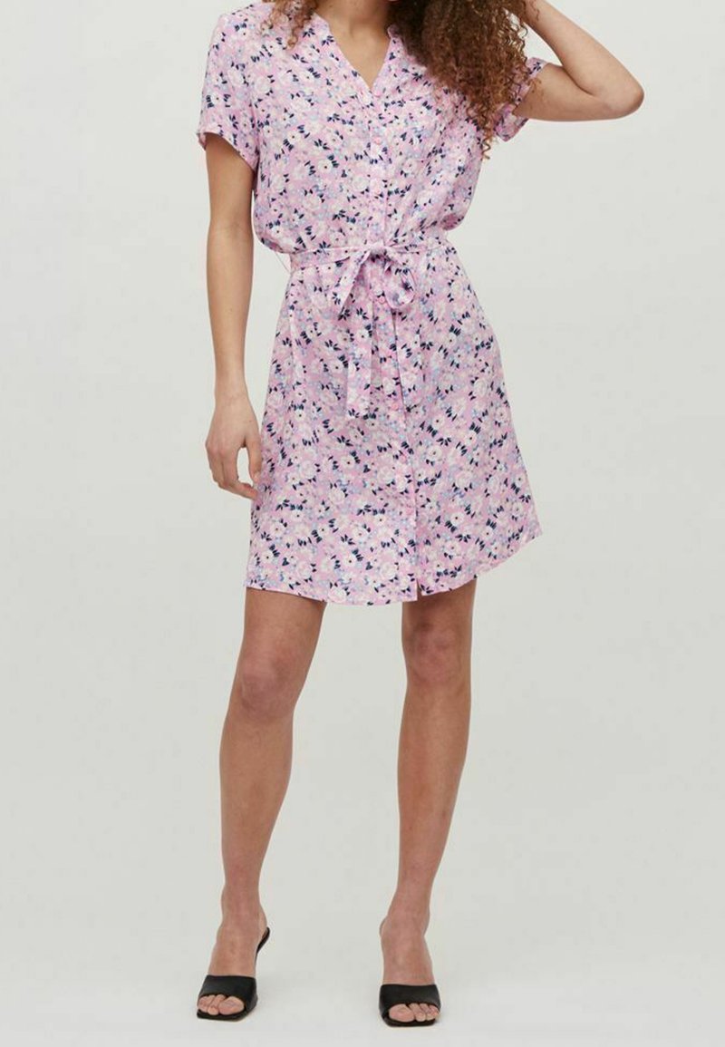 Femme portant une robe rose à fleurs à manches courtes avec des boutons, une ceinture à la taille et des talons ouverts noirs, debout avec une main posée sur sa tête.