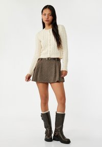 Romige kabelgebreide cardigan, gebloemde bruine rok, bruine kniehoge laarzen met romige boorden. De outfit toont een moderne gelaagde look.