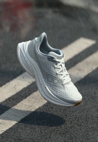 Hellgrau laufende Schuhe mit weißen Schnürsenkeln und strukturiertem Sohlendesign, die über Asphalt mit weißen Straßenmarkierungen schweben.