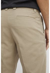 Beige Baumwollhosen mit glatter Textur, ausgestattet mit einer einzelnen Gesäßtasche und Gürtelschlaufen. Der Stoff wirkt strapazierfähig und sorgfältig verarbeitet.