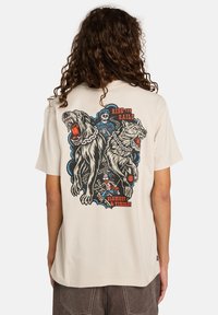 Osoba s dlouhými kudrnatými vlasy, která na sobě má béžové tričko s grafikou dvou vrčících psů na řetězech, kostry v uniformě strojvůdce a textem.