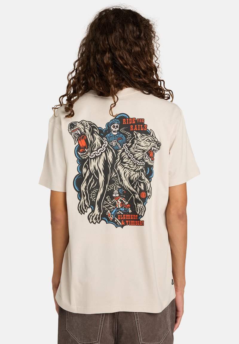 Osoba s dlouhými kudrnatými vlasy, která na sobě má béžové tričko s grafikou dvou vrčících psů na řetězech, kostry v uniformě strojvůdce a textem.
