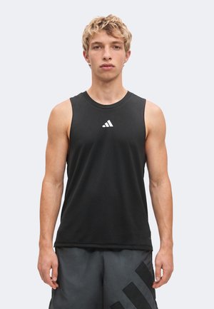 Mladý muž s krátkymi kučeravými blond vlasmi, oblečený v čiernom tielku Adidas bez rukávov a tmavých kraťasoch, stojaci pred jednofarebným pozadím.