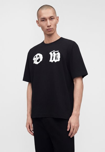 Άνδρας που φοράει μαύρο oversized t-shirt με λευκά γοτθικά γράμματα στο στήθος, στέκεται μπροστά σε μια μονόχρωμη ανοιχτογκρι φόντο.