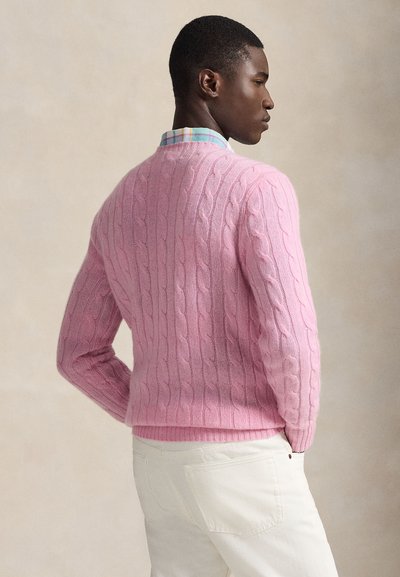 Homme portant un pull torsadé rose par-dessus une chemise à carreaux pastel et un pantalon blanc, debout de profil sur un fond neutre.
