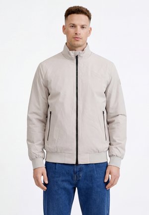 Giovane uomo che indossa una giacca leggera grigio chiaro con zip, polsini e colletto a costine, abbinata a jeans blu, in piedi davanti a uno sfondo bianco.