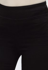 Gros plan d'une personne portant un pantalon noir ajusté et un pull en maille beige côtelé rentré à la taille.