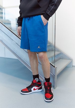 Osoba ubrana w jaskrawo niebieskie spodenki z logo Jordana, czarną bluzę z kapturem, czarne skarpetki z białym kwiatem oraz czerwono-czarno-białe buty sportowe Nike stoi przy schodach.