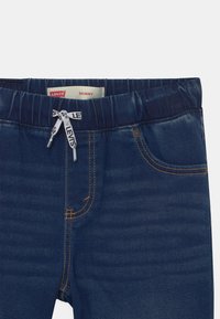 Kapeita farkut, jotka on valmistettu tummansinisestä denimestä, jossa on joustava vyötärö, vaaleanbeige ompelu, etutaskut ja brändätty nyöri.