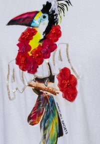 Bunter Tukan mit roten Stoffblumen und Pailletten auf weißem Stoff, der einen Ast hält, der Text "Marc Cain Sports" ist sichtbar.