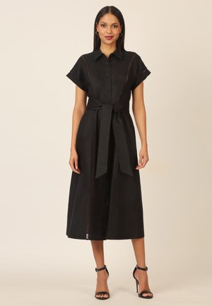 Woman standing wearing a black midi shirt dress with short sleeves, button front, waist tie, and black high-heeled sandals.