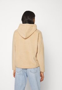 Beige fleece hoodie with a soft texture, long sleeves, and a spacious hood. The garment has a relaxed fit and a slightly curved hem.