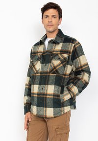 Uomo che indossa una giacca button-up a quadri verde, beige e marrone con tasche sul petto e pantaloni cargo marroni, in piedi davanti a uno sfondo bianco.