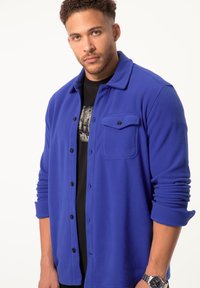 Blauwe fleeceshirt-jack met knoopsluiting, enkele borstzak en opgerolde mouwen. Draagt over een zwart graphic T-shirt.