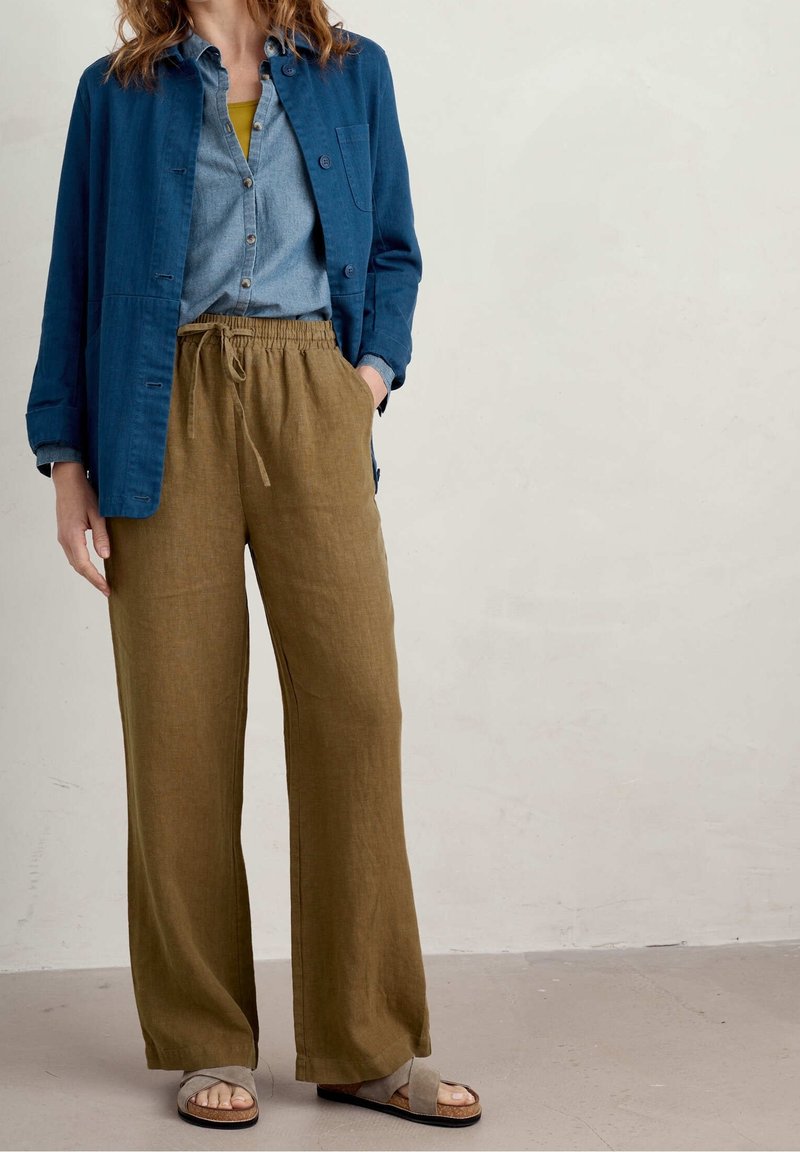 Žena v olivových lněných kalhotách na stahování, modré košili s knoflíčky vrstvené s modrou bundou a krémovými sandály, stojící před hladkou zdí.