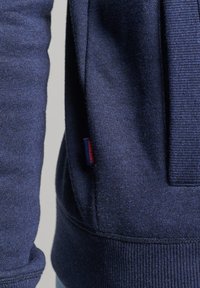 Felpa blu navy con una texture morbida, caratterizzata da un polsino a coste e da una piccola etichetta rossa. Progettata con uno stile elegante e minimalista.