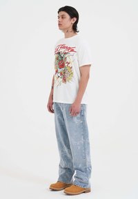 Joven con tatuajes que lleva una camiseta gráfica blanca, jeans sueltos de mezclilla azul claro con patrón y botas de trabajo color beige, de pie de perfil sobre un fondo blanco.