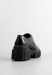 Chaussure noire en cuir brillant à lacets, avec une semelle plateforme épaisse et rainurée, et une couture blanche le long du bord de la semelle, sur une surface claire.