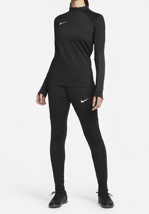 Femme debout portant une tenue de sport noire ajustée Nike avec un haut à manches longues et des leggings, avec des logos Nike blancs et des baskets noires.