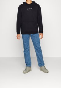 Svart hoodies med framtass, med vit broderad text, parat med ljusblå jeans och grå sneakers. Enfärgad bakgrund.