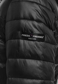 Zwarte pufferjack gemaakt van glanzend, gewatteerd materiaal. Heeft horizontale stiksels en een merklabel met de tekst "FRIEDA FREDDIES NEW YORK."