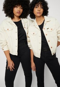 Jaquetas de ganga de creme claro com botões de cobre, dois bolsos no peito, usadas sobre tops e calças pretas. Textura suave e corte descontraído.