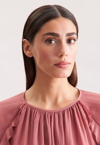 Blusa rosa pallido con pieghe e dettagli sfrangiati sul collo. Tessuto morbido con colletto rotondo, che mostra una caduta delicata.