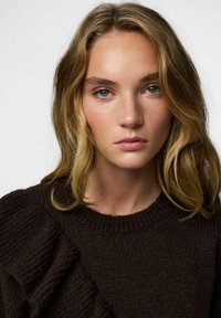 Brązowy sweter z dzianiny z prążkowanym okrągłym dekoltem i falbankowymi akcentami po jednej stronie. Gładka tekstura i dopasowany krój.